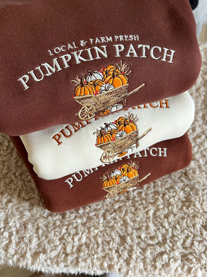 Two folded sweatshirts stacked on a textured cream rug. The top sweatshirt is dark brown with white embroidered text reading 'LOCAL & FARM FRESH PUMPKIN PATCH' and an autumn pumpkin wheelbarrow graphic.