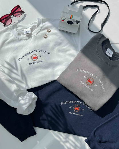 A photograph of three sweatshirts in White, Charcoal and Navy all bearing a Fisherman's Wharf inspired design. A pair of sunglasses, a Polaroid camera and some earrings are scattered for decoration.